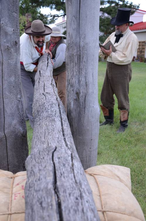 Three men in historical attire work together to lift a large log, while another man observes and takes notes.