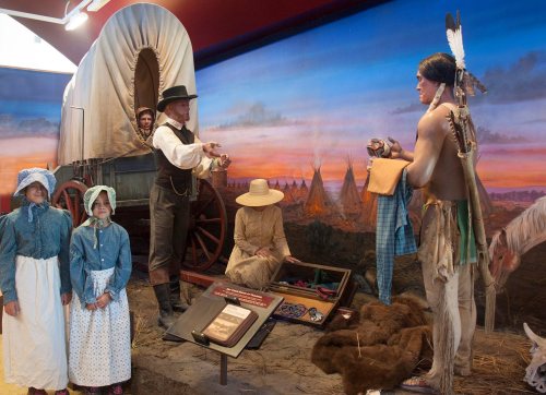 A historical scene depicting a Native American trading with settlers near a covered wagon, with children observing.