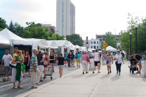 A bustling outdoor market with people walking among white tents and vendors, surrounded by trees and buildings.