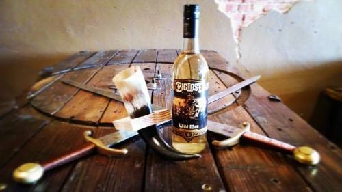 A bottle of Big Lost Wild Man liquor surrounded by swords and a horn on a wooden table.