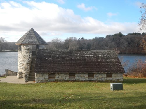 A stone building with a tower by a lake, surrounded by trees and grass under a partly cloudy sky.