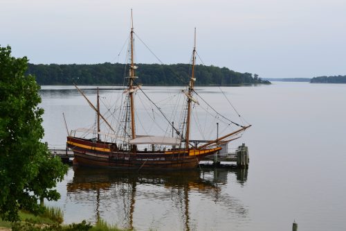 A historic wooden ship docked by a calm river, surrounded by trees and a serene landscape.
