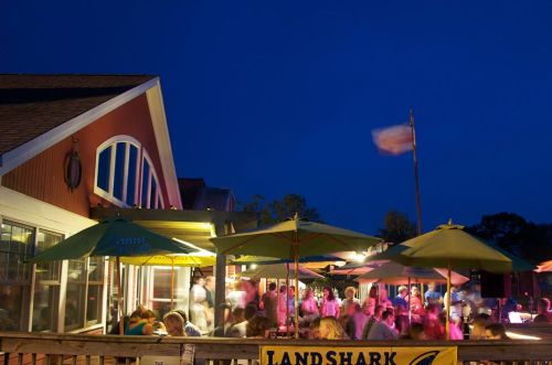 A lively outdoor restaurant scene at night, with people dining under umbrellas and a flag waving in the background.