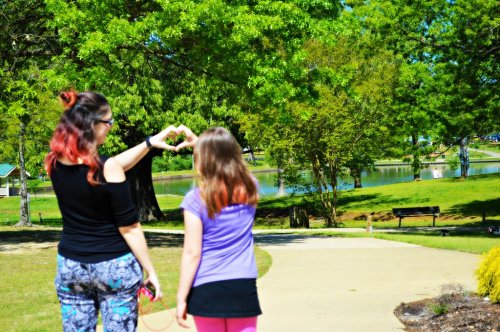 Two people in a park making a heart shape with their hands, surrounded by greenery and a serene lake in the background.