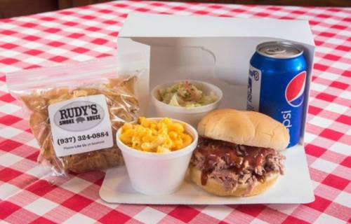 A takeout meal featuring a pulled pork sandwich, macaroni and cheese, coleslaw, chips, and a can of soda on a checkered tablecloth.