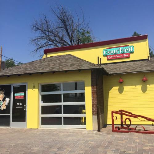 Bright yellow building with a sign reading "GourMelt Grilled Cheese Shop," featuring large windows and a welcoming entrance.