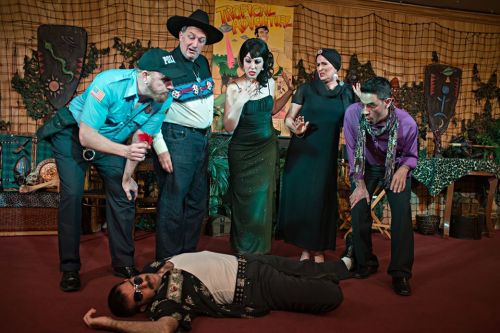 A theatrical scene with five actors in costumes, gathered around a man lying on the ground, set in a jungle-themed backdrop.