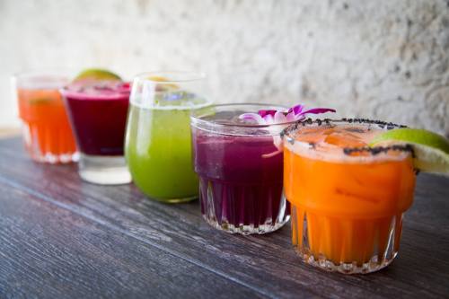 A colorful array of five cocktails in glasses, featuring vibrant hues of orange, green, purple, and red, garnished with lime.