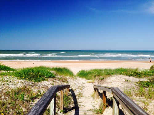 A wooden walkway leads to a sandy beach with gentle waves under a clear blue sky.