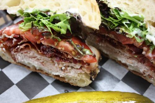 Close-up of a sandwich with layers of meat, lettuce, and tomato, served with a pickle on the side.