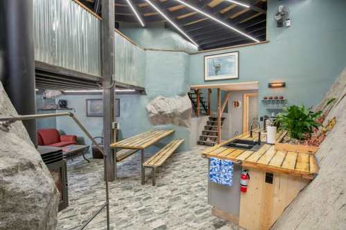 Interior of a modern space featuring rock walls, wooden tables, a kitchen area, and a staircase. Brightly lit and stylish.