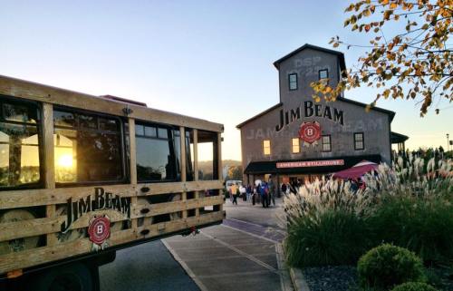 A wooden Jim Beam tour bus parked outside the Jim Beam distillery at sunset, with people gathered nearby.