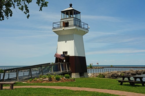 A white and brown lighthouse stands by the water, with a grassy area and benches in the foreground under a clear blue sky.
