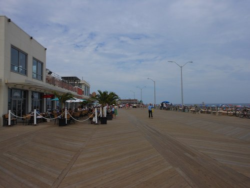 A wooden boardwalk lined with cafes and umbrellas, leading to the beach under a cloudy sky.
