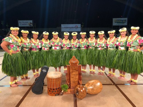 A group of women in traditional Hawaiian attire, adorned with flowers, posing together with musical instruments.
