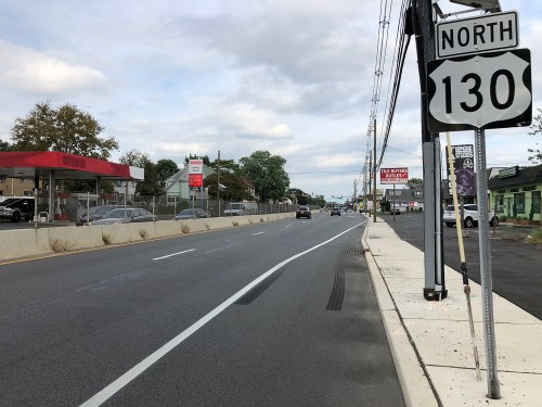 A view of a road with a "Route 130 North" sign, featuring cars, businesses, and cloudy skies in the background.
