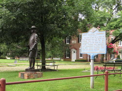 A bronze statue of a man stands in a park, surrounded by trees and a historical sign about New Castle Common.