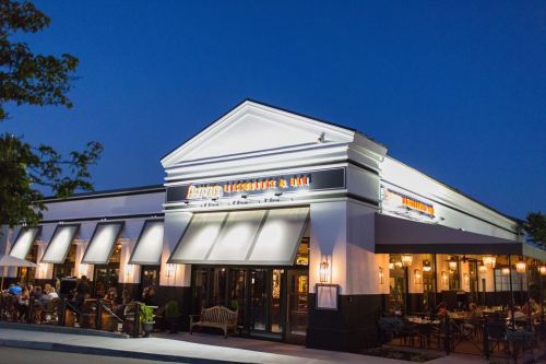 A well-lit restaurant and bar exterior at night, featuring outdoor seating and large awning-covered windows.
