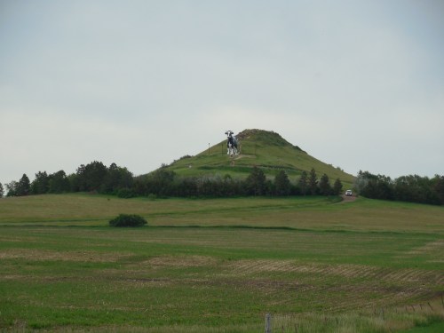A large cow statue atop a grassy hill, surrounded by fields and trees under a cloudy sky.