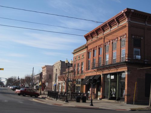 Historic brick buildings line a street under a clear blue sky, with outdoor seating and parked cars visible.