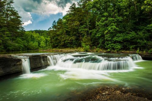 A serene waterfall cascades over rocks into a tranquil green river, surrounded by lush trees and a cloudy sky.