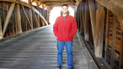 A person in a red jacket stands on a wooden bridge, smiling, with light shining in the background.