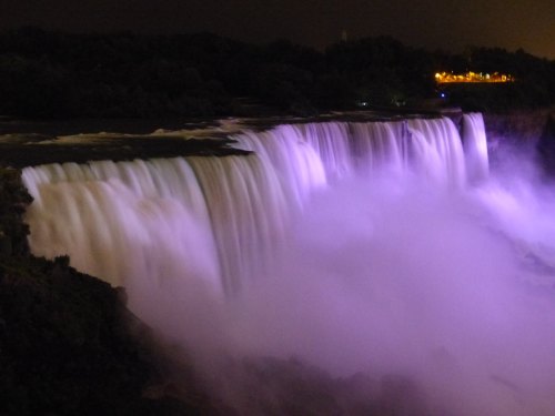 A nighttime view of a waterfall illuminated in purple, surrounded by dark foliage and distant lights.