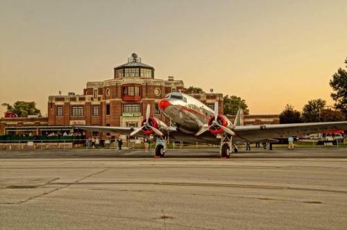 A vintage airplane parked in front of a historic building at sunset, with people gathered nearby.