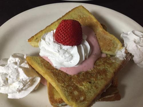 A plate of French toast topped with whipped cream and a strawberry, served with a dollop of pink cream.