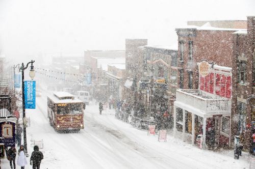 A snowy street scene with a trolley, shops, and people walking in a winter storm.