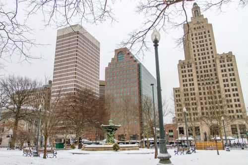 Cityscape featuring tall buildings and a fountain in a snowy park, with bare trees and a cloudy sky.