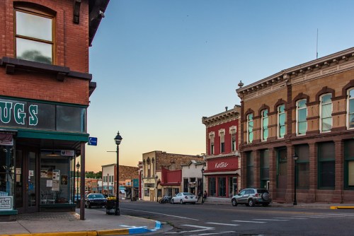 Historic downtown street scene with brick buildings, shops, and parked cars under a clear sky at dusk.