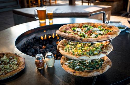 Three stacked pizzas on a table near a fire pit, with two beers and condiments in the background.