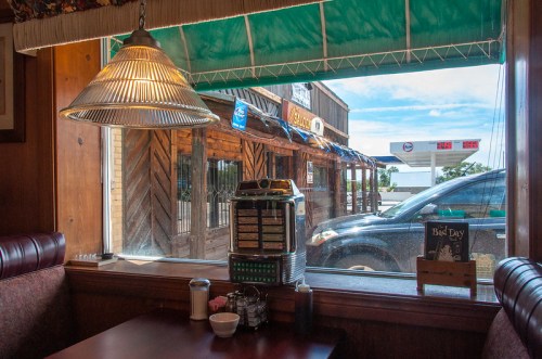 A cozy diner interior with a jukebox, looking out at a street with a car and rustic buildings.