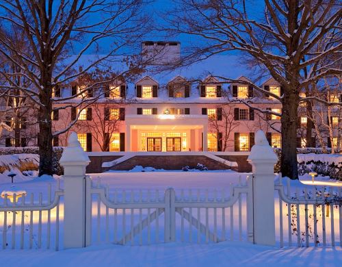 A grand house illuminated at dusk, surrounded by snow-covered trees and a white picket fence.