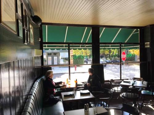 Two people sit at a table in a cozy café, looking out through large windows with green awnings.
