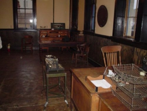 An old, empty room with wooden furniture, including a desk, chairs, and a vintage typewriter on a small table.