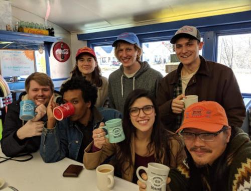 A group of seven friends in a diner, smiling and holding coffee mugs, wearing colorful hats.