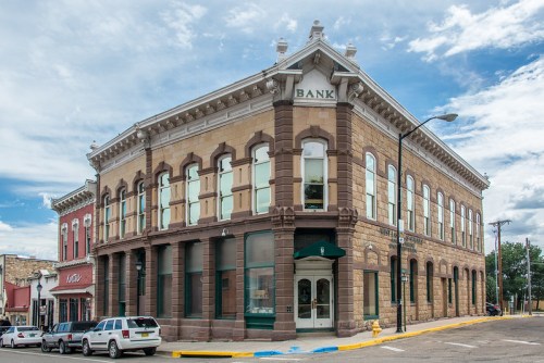 Historic bank building with large windows, stone facade, and a blue sky in the background. Cars parked nearby.