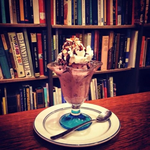 A dessert sundae in a glass dish topped with whipped cream, chocolate drizzle, and almonds, set against a bookshelf.