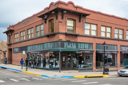 Historic brick building with "Plaza Drugs" sign, featuring large windows and pedestrians outside on a cloudy day.