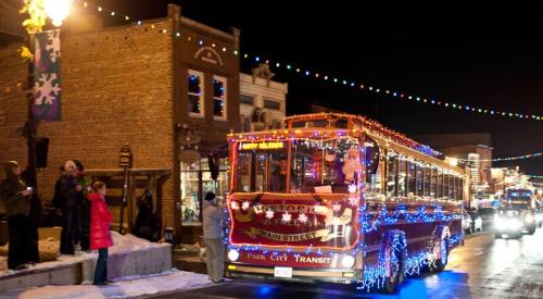 A festive trolley adorned with colorful lights drives through a snowy street, surrounded by holiday decorations and onlookers.