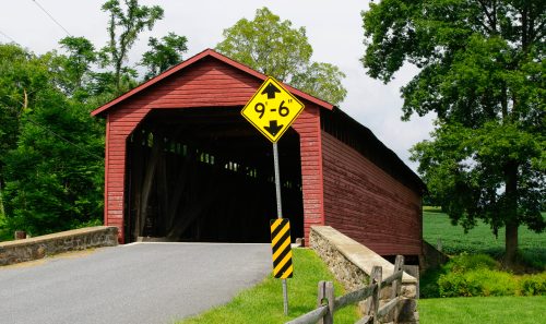A red covered bridge with a low clearance sign, surrounded by trees and a rural landscape.