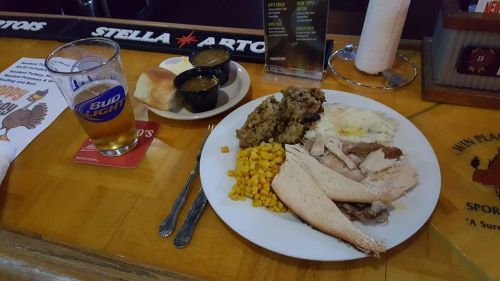 A plate of turkey, stuffing, corn, and mashed potatoes, with a side of rolls and gravy, next to a beer on a bar counter.
