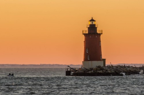 A red lighthouse stands on a rocky shore at sunset, with a small boat in the foreground and calm waters.