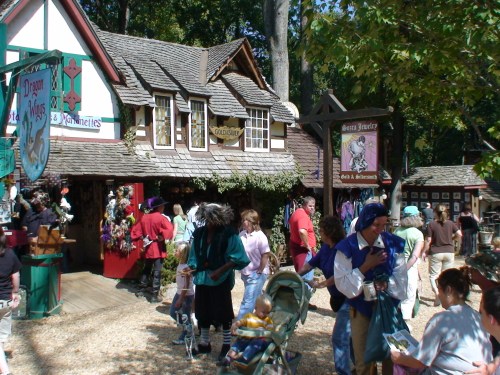 A bustling outdoor market with people browsing shops, colorful costumes, and various stalls in a wooded setting.
