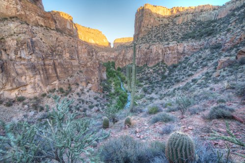 A scenic canyon landscape with steep rock walls, a winding river, and desert vegetation under a clear sky.