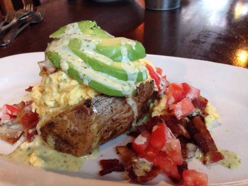 A hearty dish featuring scrambled eggs on toasted bread, topped with avocado, diced tomatoes, and a creamy sauce.