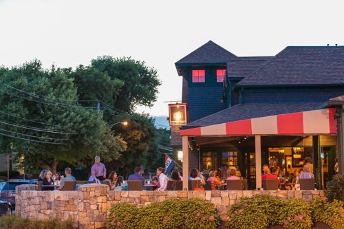 A lively restaurant patio filled with people, surrounded by greenery and a charming building at dusk.
