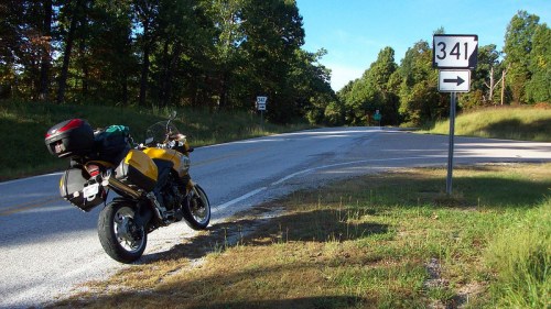 A yellow motorcycle parked beside a rural road with a directional sign for Route 341 and trees in the background.